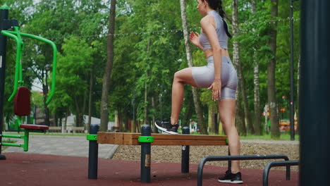 A-young-woman-in-a-Park-performs-striding-lunges-on-a-bench-in-sportswear-in-the-summer.-Athletics-Caucasian-woman-trains-in-the-Park.