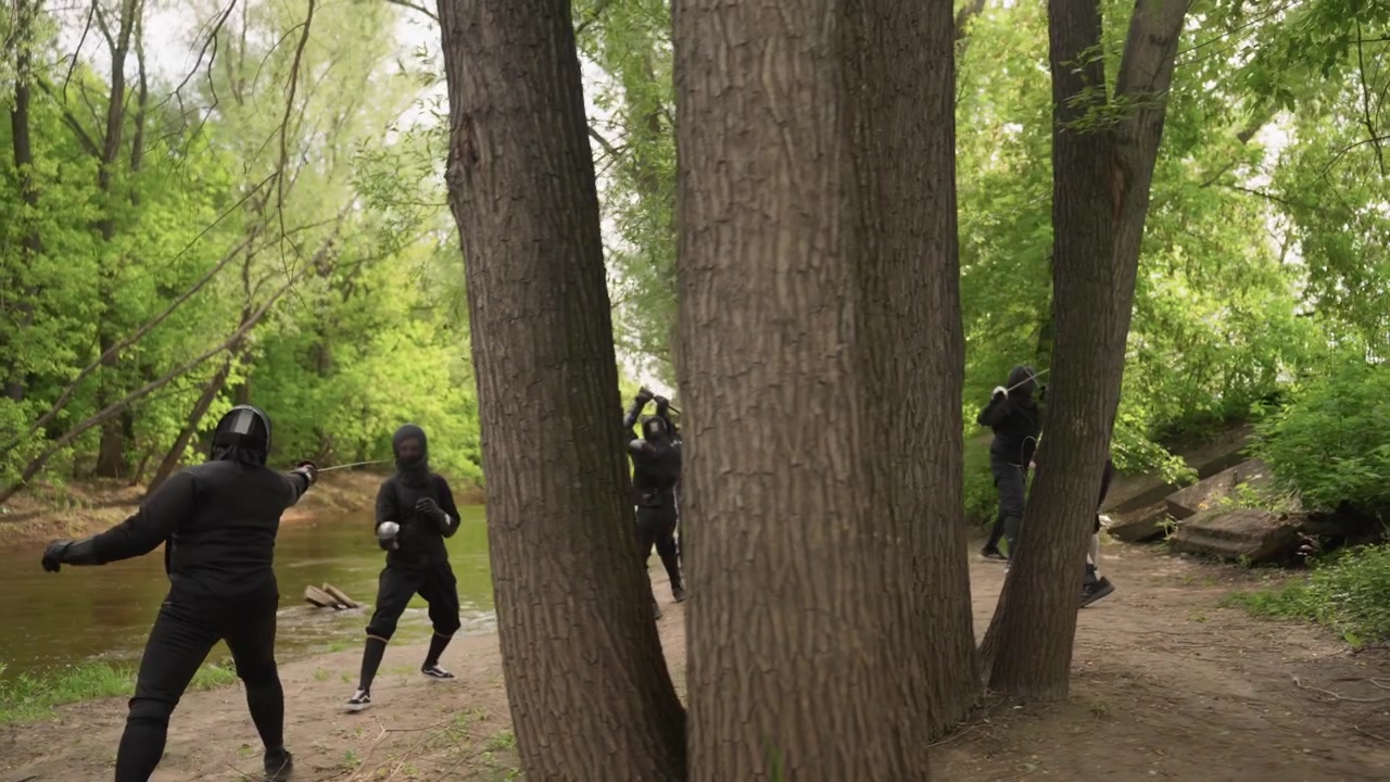 Multiple people dressed in black protective gear engage in coordinated movement along forest path near water demonstrating tactical maneuvers and weapon handling. scene features dense green