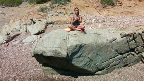 mujer caucásica meditando en una roca junto a la costa del mar en cerdeña, italia en un día soleado