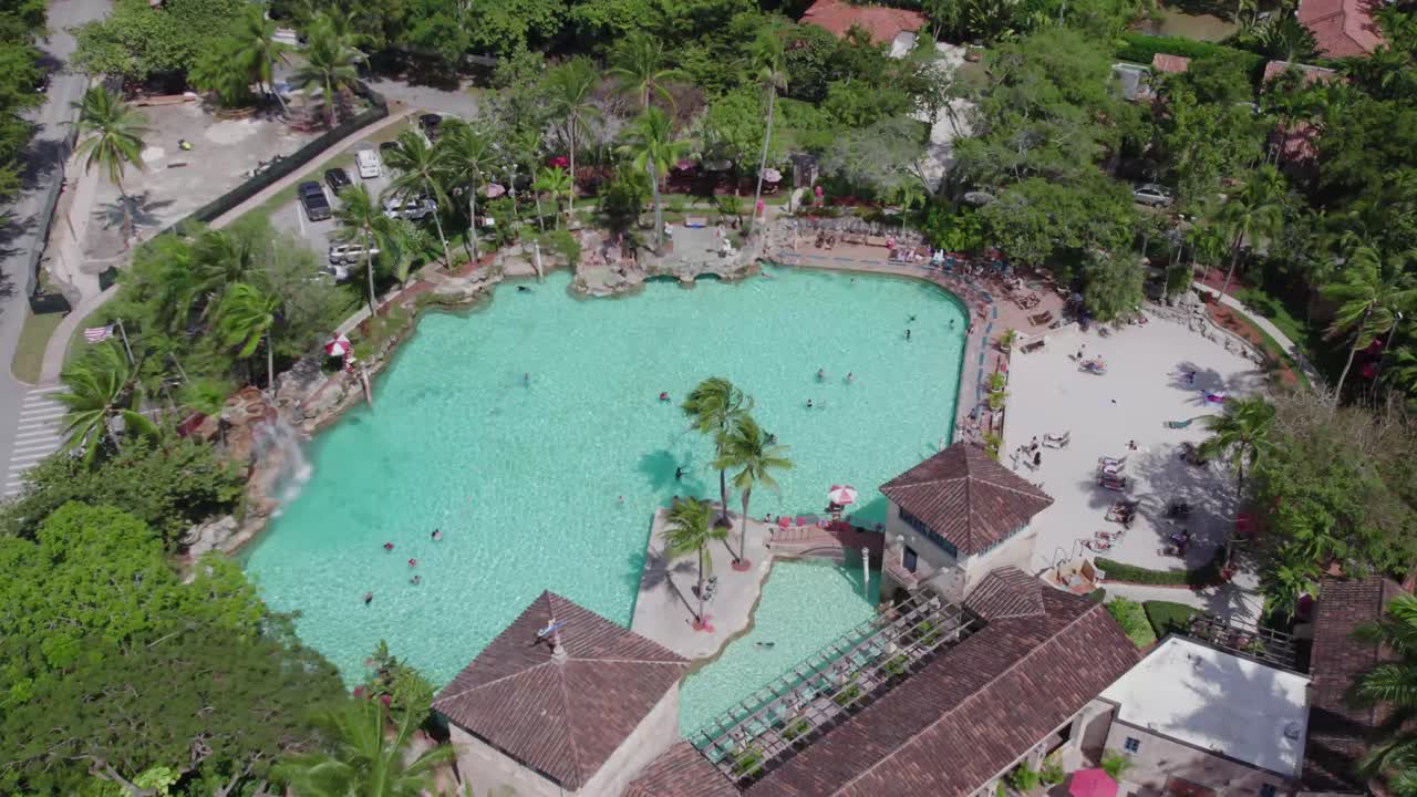 Deslize suave de drone sobre a Venetian Pool ensolarada, exibindo sua enorme lagoa e paisagem tropical.