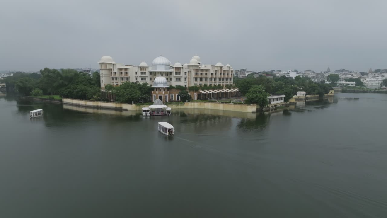 Un bateau de passagers à toit blanc approche d'un complexe hôtelier de style palais orné sur le rivage du lac. Un ciel couvert projette une lumière douce sur l'eau, les terrasses murées et les dômes d'inspiration moghole entourés d'une verdure luxuriante.