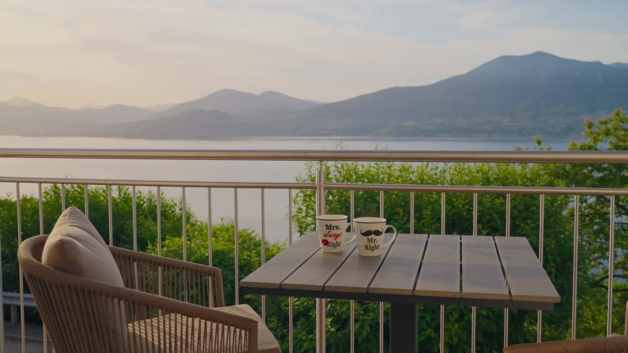 Inquadratura statica di un balcone tranquillo con una sedia e due tazze su un tavolo che si affaccia su uno splendido lago di montagna al tramonto, con una luce calda e soffusa che illumina le cime