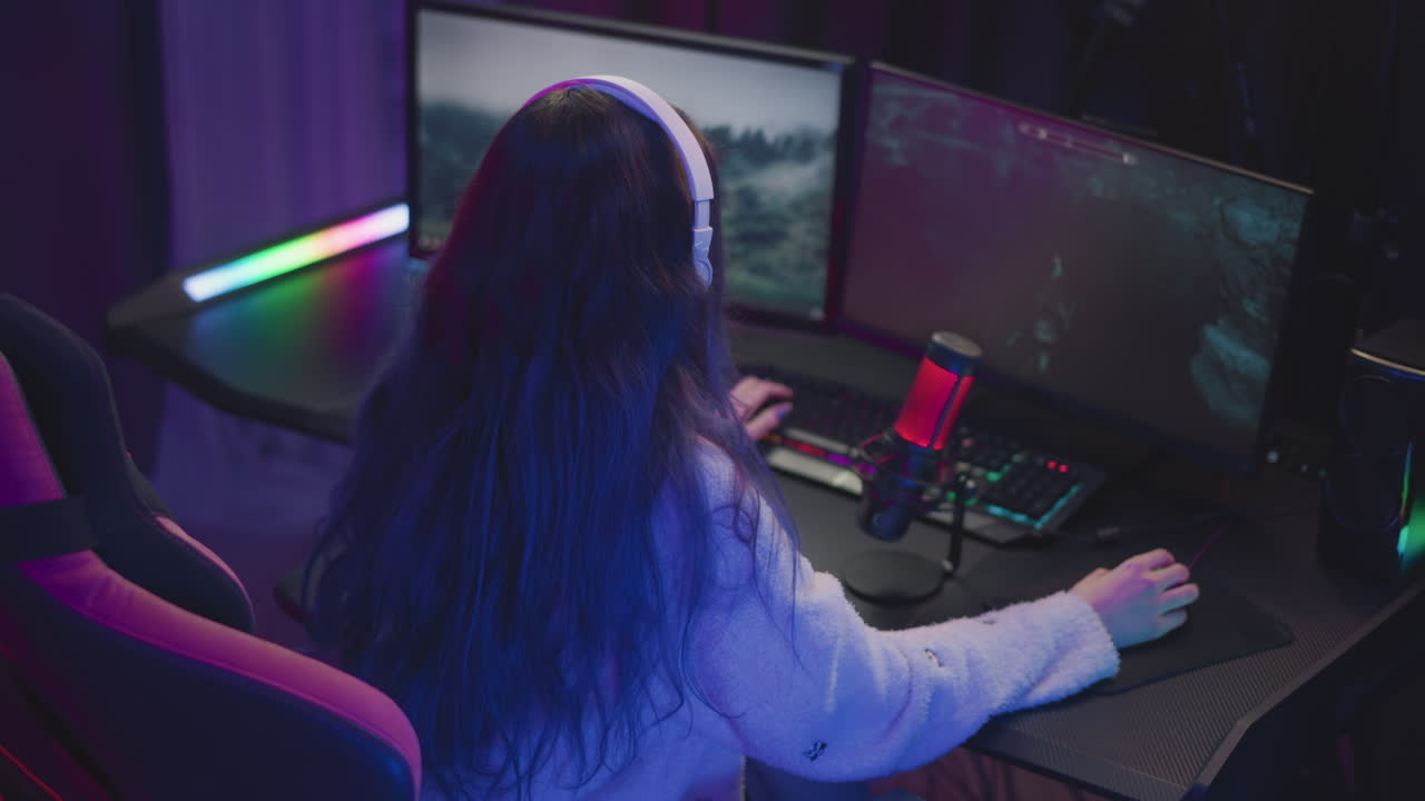 back view of female gamer wearing white headset and fluffy sweater playing immersive video game on dual monitors with rgb lighting, mechanical keyboard, and red microphone in dark gaming room setup