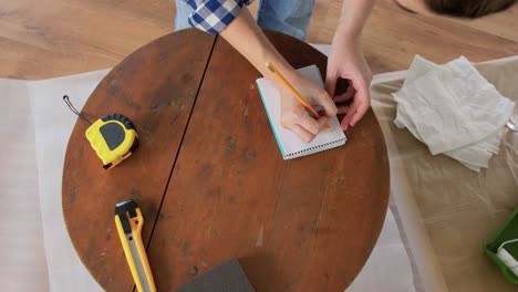 Woman-with-Ruler-Measuring-Table-for-Renovation.repair,-diy-and-home-improvement-concept--woman-with-ruler-measuring-old-round-wooden-table-for-renovation-and-writing-to-notebook