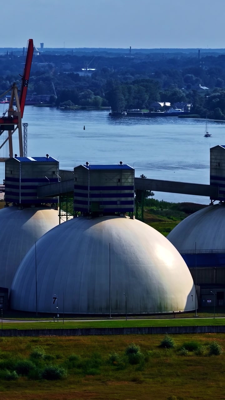 Grandi cupole di stoccaggio industriali si trovano vicino a un fiume calmo, con una barca a vela visibile in lontananza. Ideale per documentari ambientali o servizi di design industriale.