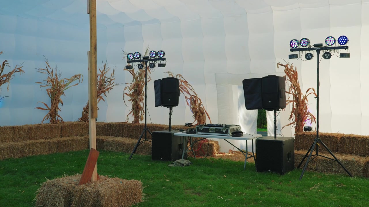 Rustic outdoor event setup with DJ equipment, speakers, lighting, hay bales, and corn stalks in a bright inflatable tent.