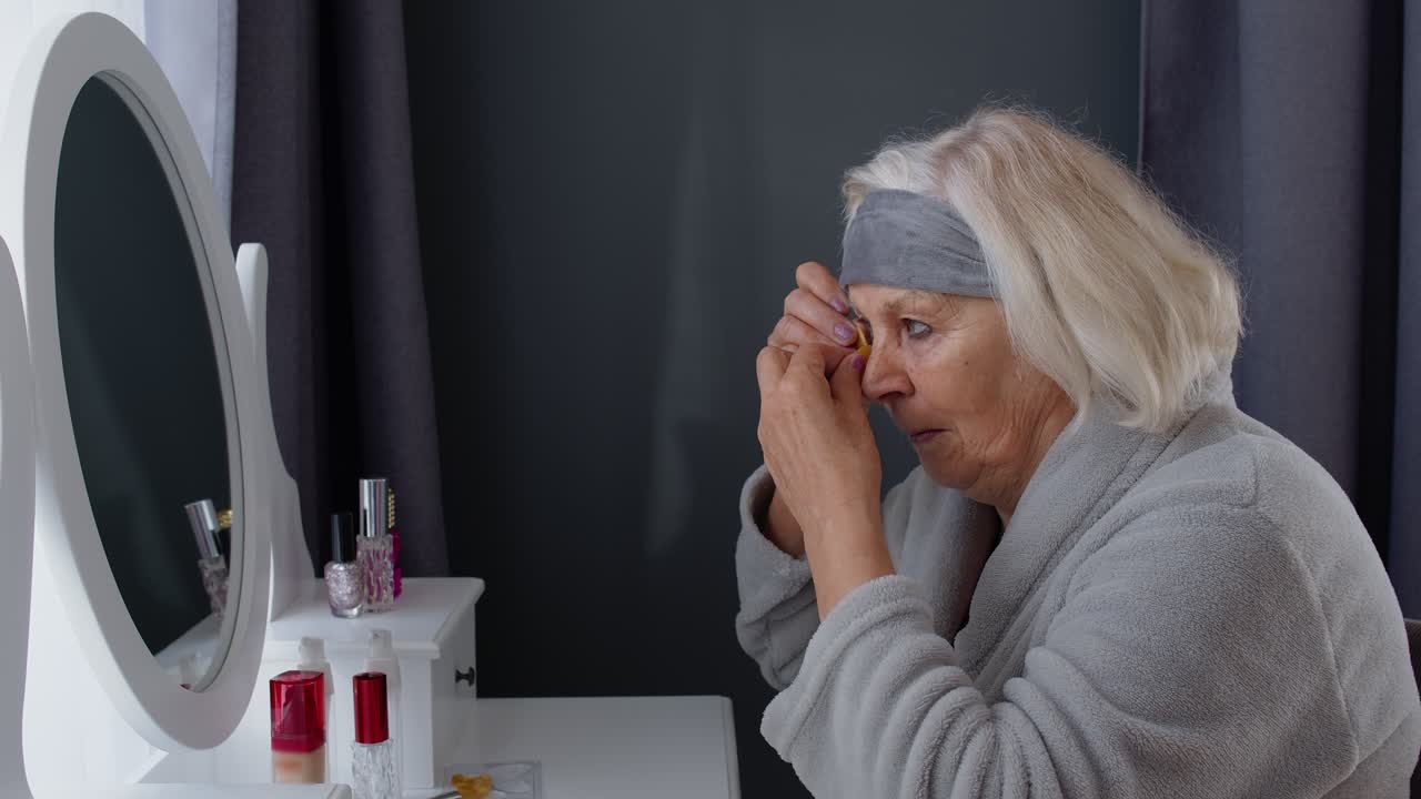 Old senior elderly woman grandmother applying anti-wrinkle eye patch, putting makeup on at home