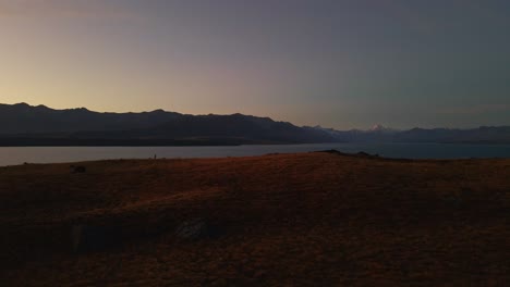 Tiny-man-on-ridge-line-above-massive-lake,-snow-covered-mountains-in-background