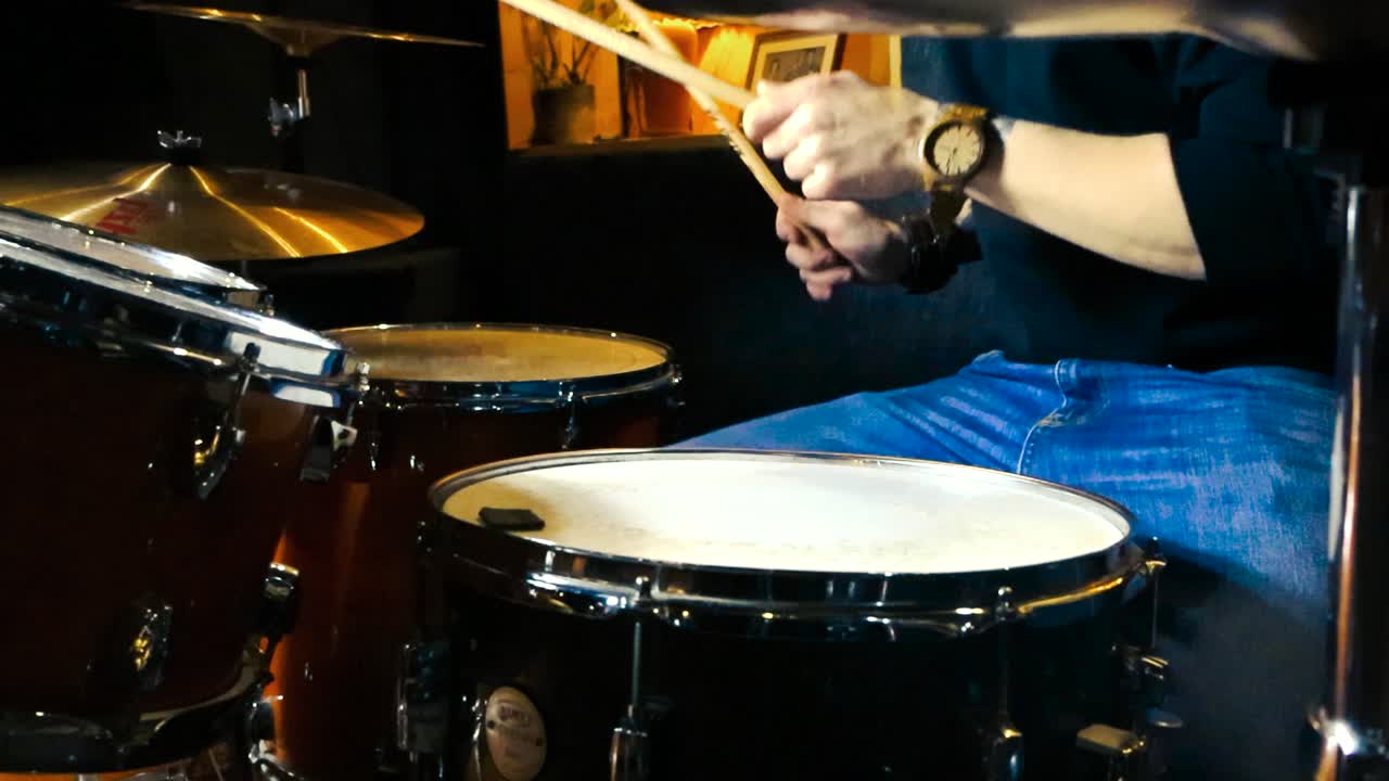 Lateral view of man playing drums in dimly lit studio room. Musician hands with drumsticks beating drums and strike the cymbals. Soft glow on drum set. Drummer beating rhythm, scenic drumming angles