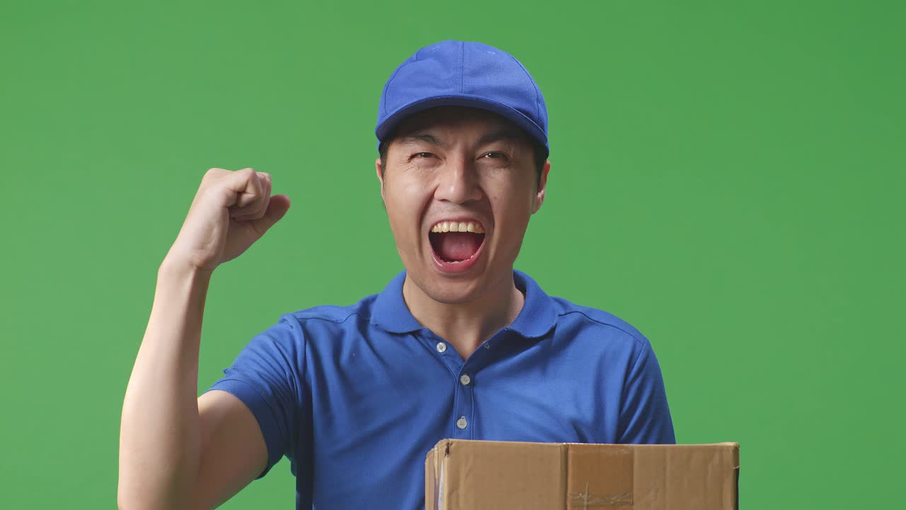 푸른 유니폼을 입은 아시아 남성 택배사가 성공을 축하하며 스튜디오의 녹색 스크린 배경에 박스를 배달합니다.