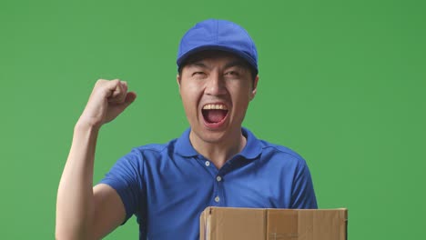 primer plano de un mensajero asiático en uniforme azul celebrando el éxito entregando una caja de cartón en el fondo de la pantalla verde en el estudio