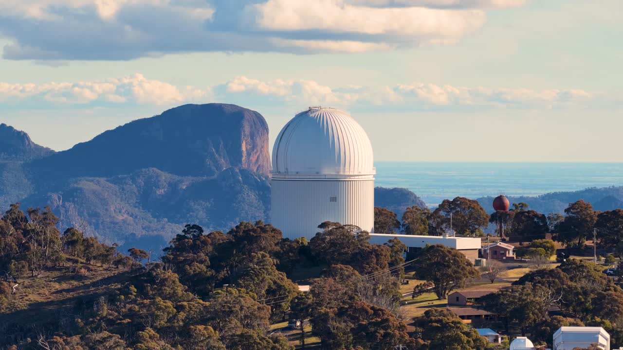 Aerial footage circles a white observatory dome atop a forested mountain ridge, with dramatic peaks and soft sunset lighting in the background