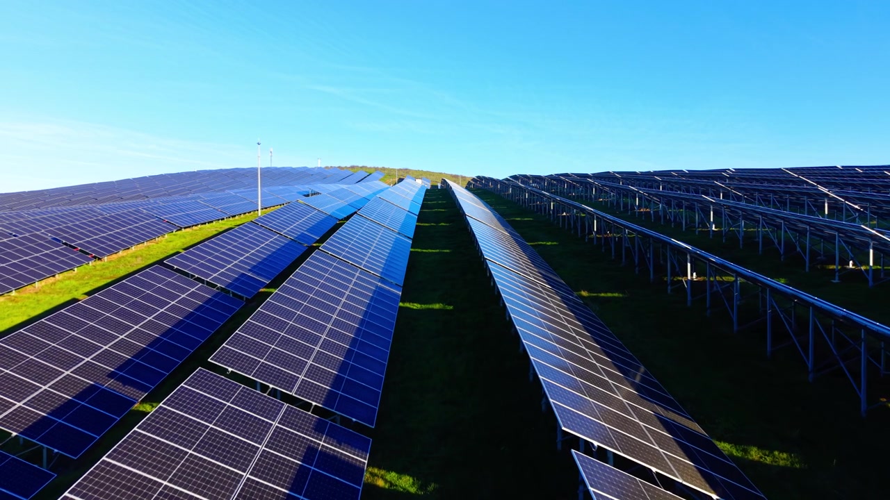 Longa fileira de painéis solares sob céu azul claro. Imagem de drone mostrando painéis se estendendo por uma encosta ensolarada.