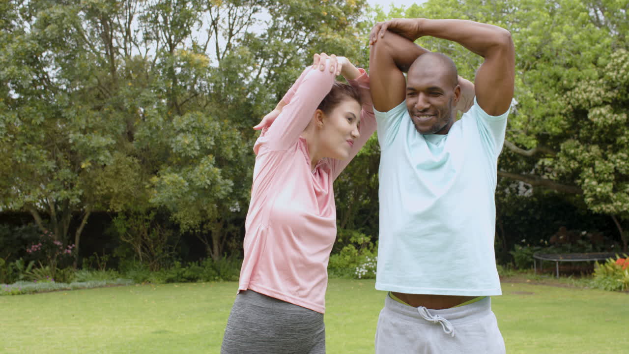 Diverse multiracial couple stretching arms outdoors, smiling and enjoying exercise, in garden