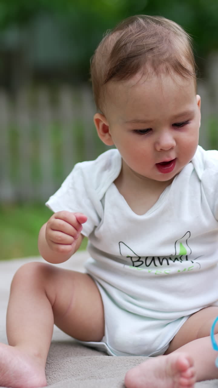 Tiny toddler sitting outdoors playing with his bottle of water. Lovely baby tries to change position to crawl ahead. Blurred backdrop. Vertical video