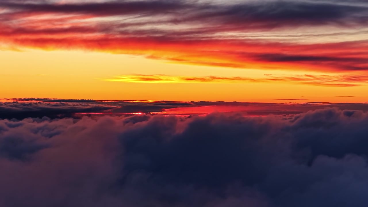 Atemberaubende Luftaufnahme über einem dichten Wolkenmeer während eines dramatischen Sonnenuntergangs. Zeigt leuchtende Bänder aus orangefarbenem, gelbem und rotem Licht, die sich über einen weiten Himmelshorizont ziehen.
