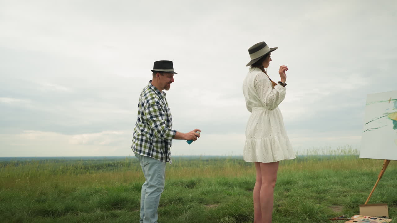 мужчина в клетчатой рубашке осторожно распыляет репелент от комаров на спине женщины, когда она носит стильную шляпу и белое платье. они находятся в пышном травяном поле с частично окрашенным полотном поблизости