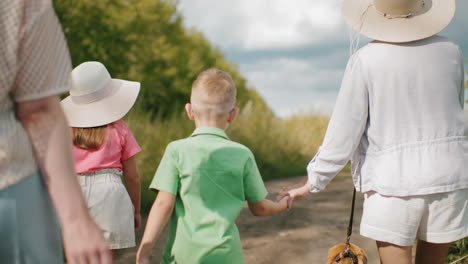 woman in white hat holding son hand while girl in pink walks alongside on dirt path, surrounded by greenery under partly cloudy sky, with partial view of another person carrying basket behind them