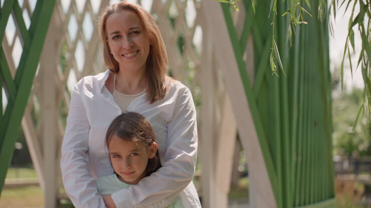 Mutter und Tochter umarmen sich innig an einem Pavillon, die Mutter lächelt, während das Kind sich anlehnt, sanftes Sonnenlicht und Gitterhintergrund, ruhige, liebevolle Stimmung, zärtliche, fürsorgliche Interaktion an einem Nachmittag im Freien.