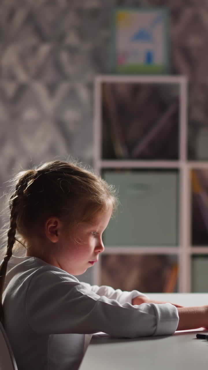 Upset little girl with braid draws picture on paper sheet with green crayon sitting at table in children room close side view slow motion