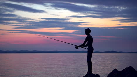silhouette of a shirtless young man standing on the rocks near sea while trying to catch a fish during sunset - wide shot