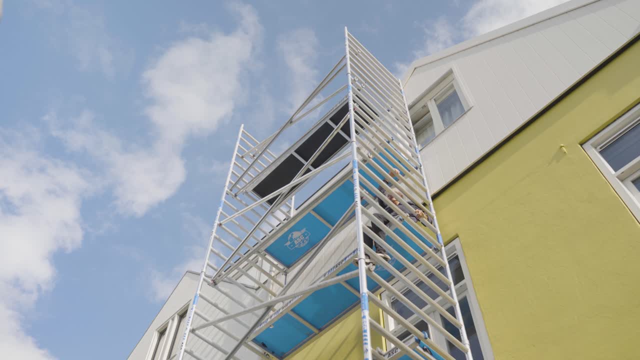 Close-up shot of an elderly woman climbing scaffolding to reach the wall of her house, representing persistence, vitality, and determination to remain independent