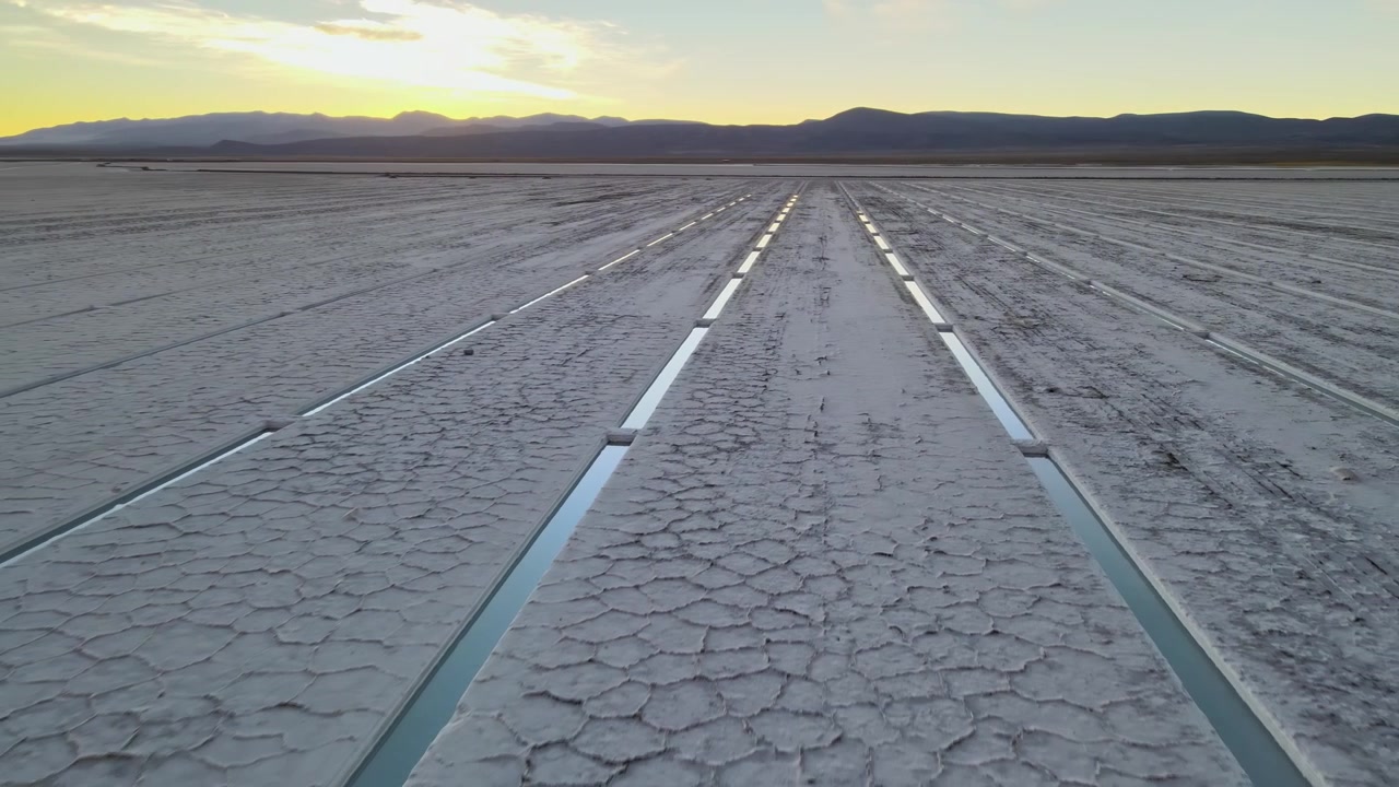 Parallel lithium extraction trenches cut across the cracked white surface of Salinas Grandes at sunrise, with distant mountains framing the high-altitude salt flat.