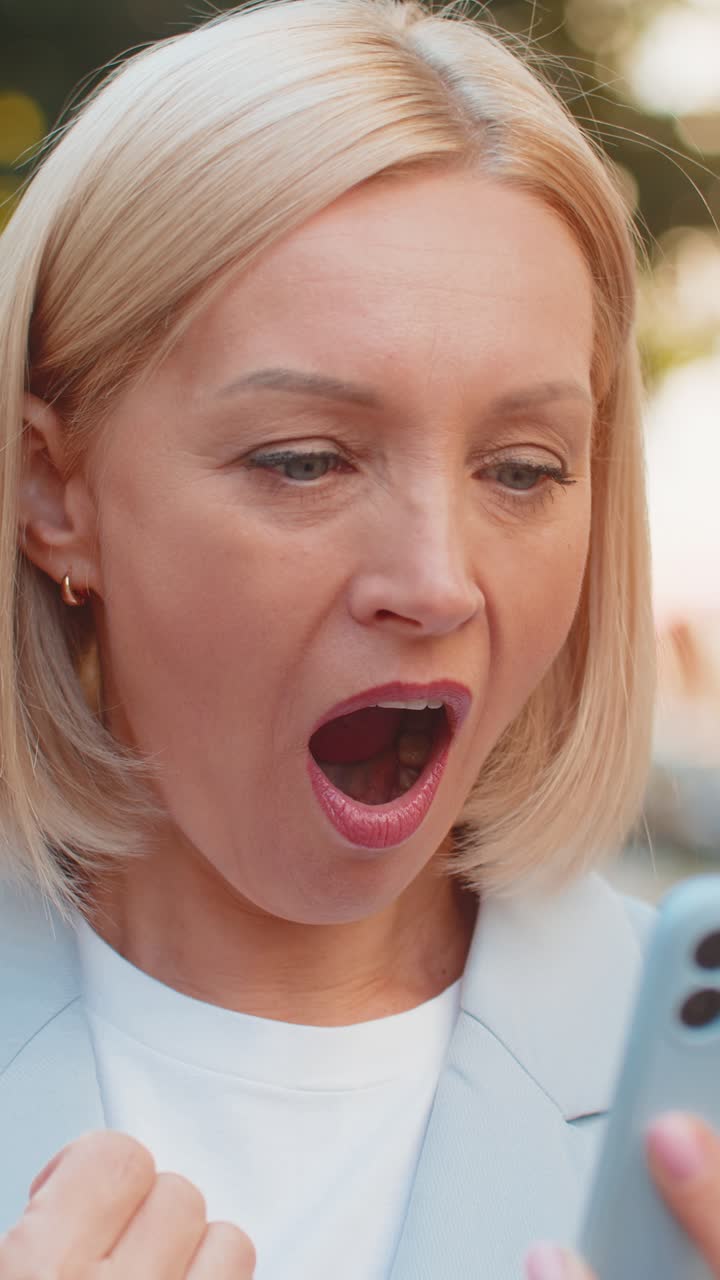 Caucasian mature businesswoman looking shocked while reading good news on smartphone on city street