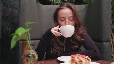 young woman with long brown hair sits in cozy booth, gently sipping latte from white cup with eyes closed. table in front shows creamy waffles with strawberries and small decorative plant in vase
