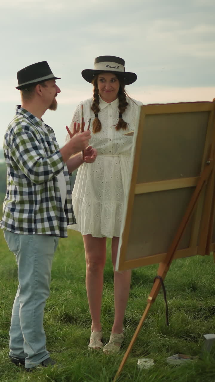 An artist in a black hat and checkered shirt hands a paint palette to a woman in a white dress. They stand together in a serene grassy field overlooking a calm lake under a cloudy sky