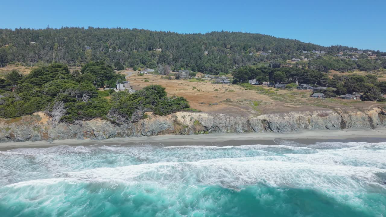 A high drone shot moves from the ocean toward Black Point Beach, showing cliffs, sandy shore, coastal houses, and the forest beyond