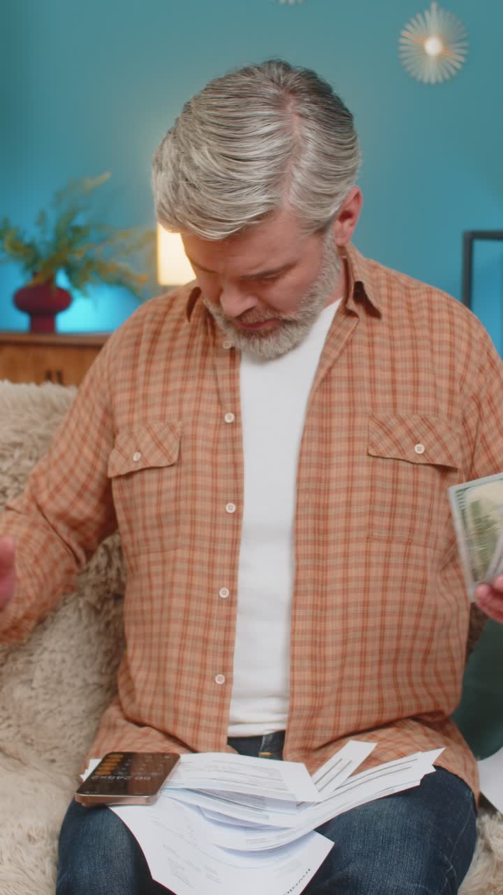 Mature man on sofa using smartphone calculator counting cash and bills feeling exhausted at home