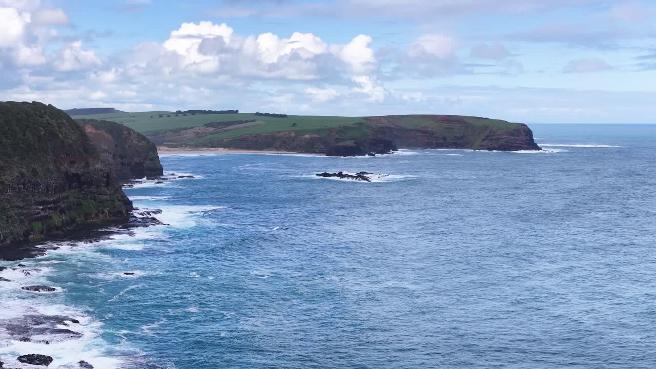 Drone camera glides above rugged coastline, capturing waves, cliffs, and blue sky in daylight