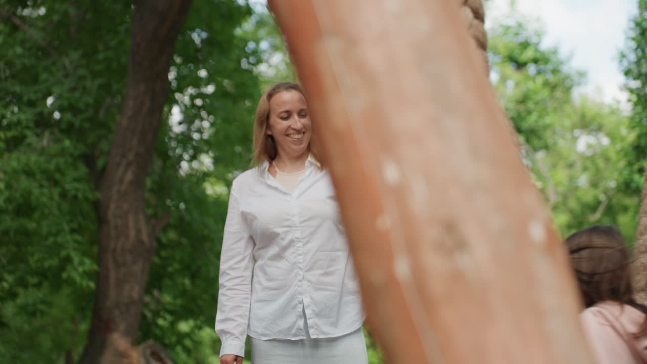 Madre e figlia in un parco alberato, la mamma aiuta la bambina a salire su una grande altalena di corda intrecciata, sorriso caloroso, camicia bianca casual, luce estiva, insegnamento delicato, stretta supervisione, famiglia spontanea.