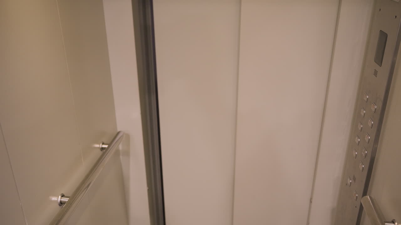 Empty elevator opening to dim hallway, metal handrail and beige panels, carpeted corridor beyond, soft overhead light, slow reveal of entrance, quiet domestic building, minimal composition, waiting