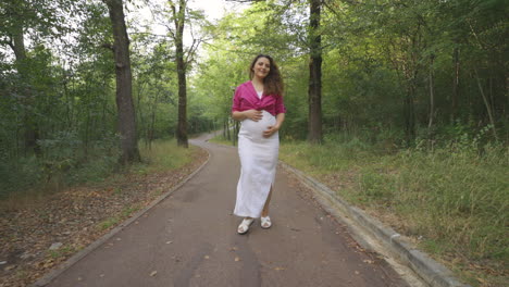 happy pregnant woman expectant mother walking alone in nature, attractive curly casually dressed mom turning around stroking caressing touching her belly smiling, childbearing love, followed by camera