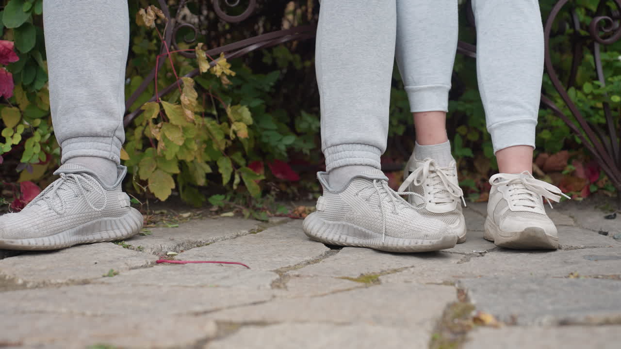 Leg view of two people seated outdoors in matching light gray outfits and sneakers, standing on stone pavement with green and red autumn leaves swaying gently