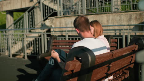 adults studying outdoors on bench; woman shows something on device to man, tranquil urban backdrop with iron railings and staircase illuminated by sunlight