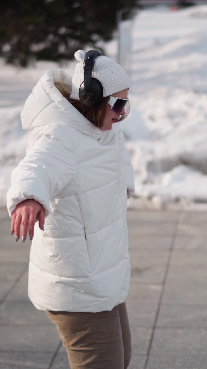 Woman wearing white puffer jacket beige pants and black headphones dances outdoors on snow covered ground. Her movements are fluid with arms extended or bent at elbows set against urban