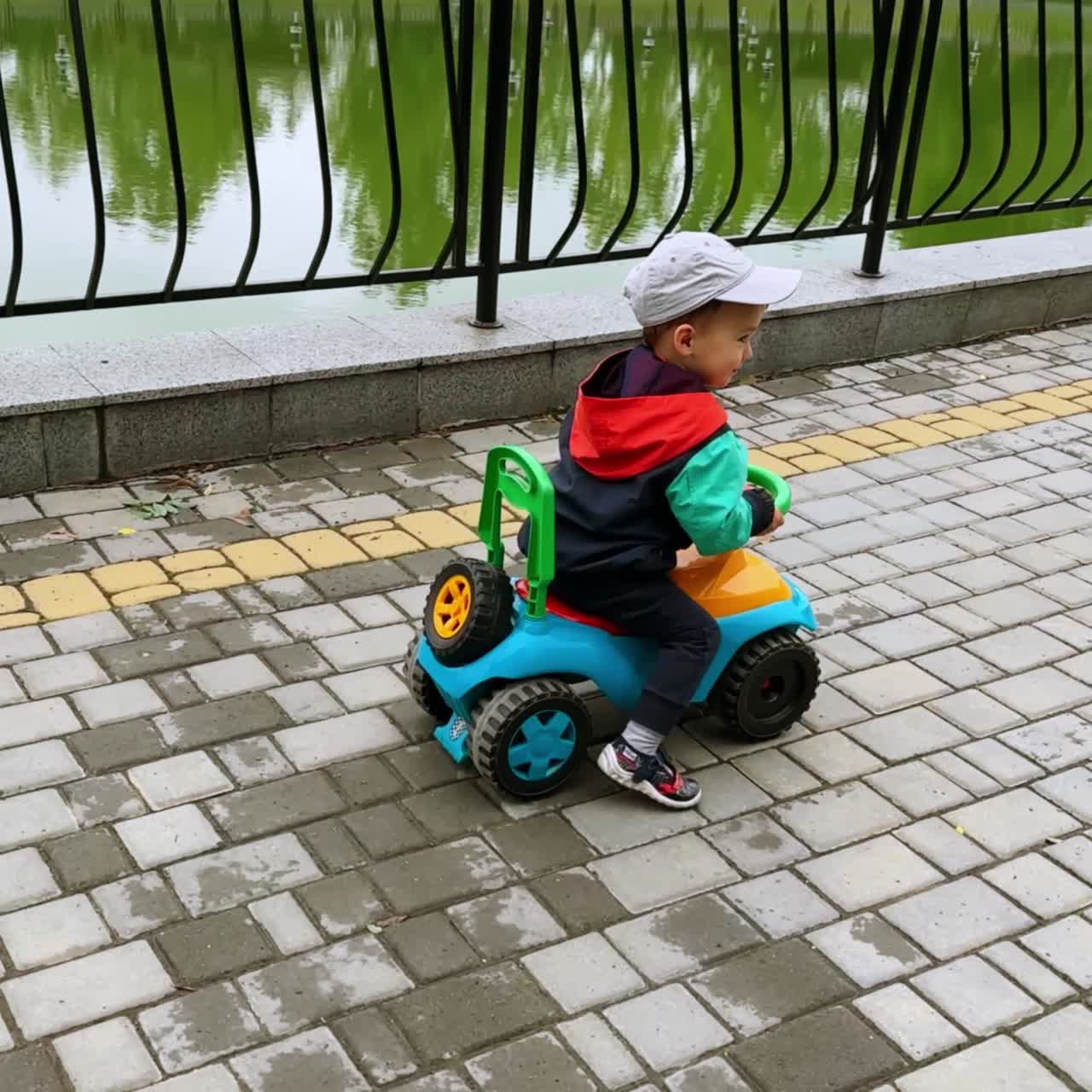 Adorabile bambino che guida la sua macchinina giocattolo spingendosi con i piedi. Il bambino gioca lungo il laghetto artificiale.