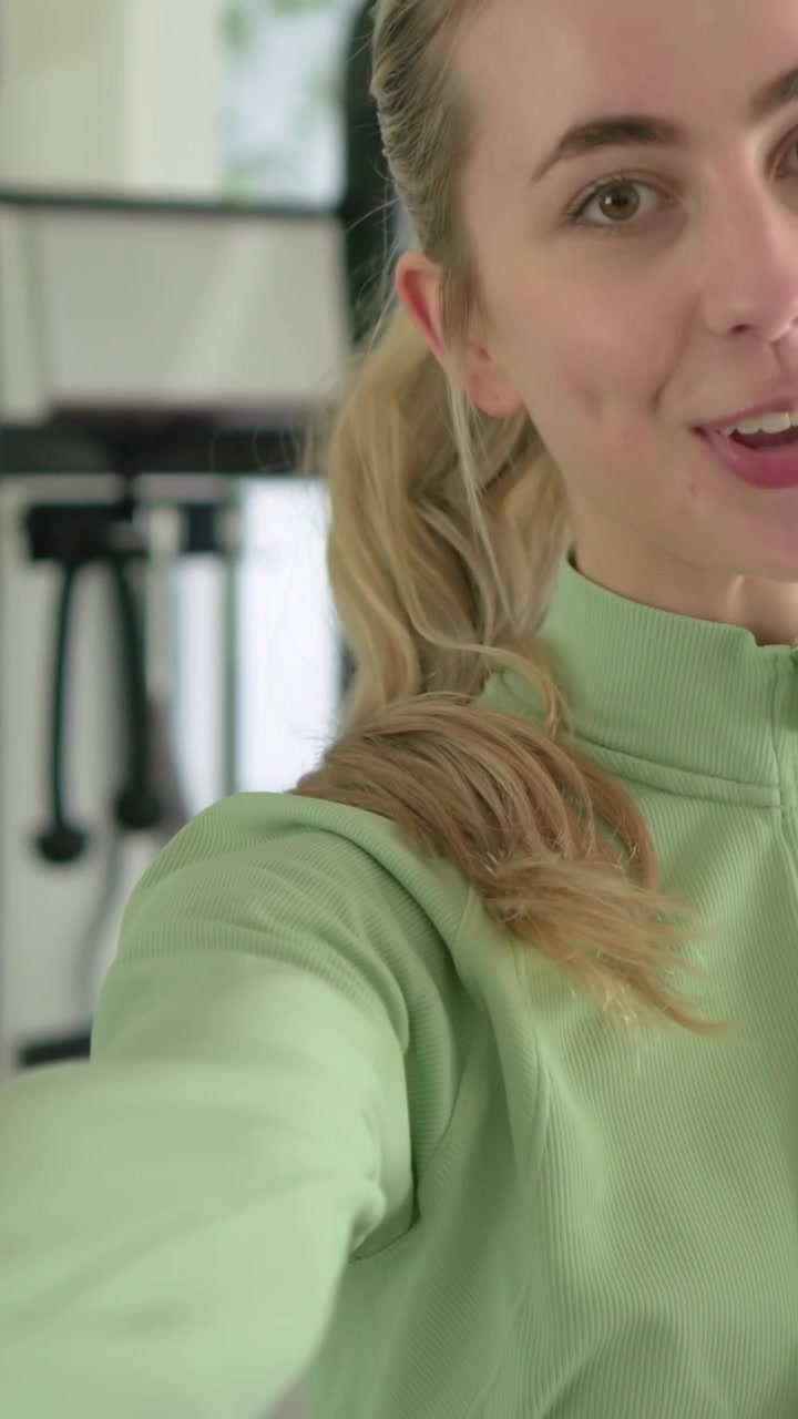 Video verticale: Donna in top verde che saluta mentre registra e allunga la mano verso la telecamera su una macchina da palestra a casa.