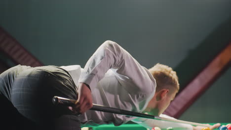 close-up back shot of player in white shirt and grey trousers leaning over green pool table, gripping cue stick with precision. overhead lights cast glow strike in dimly lit billiard hall