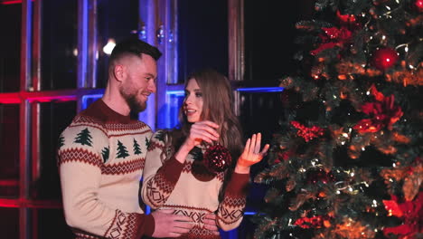 happy couple decorating christmas tree at home. beautiful woman and her husband in the same knitted christmas sweaters hang christmas ball on new year tree.
