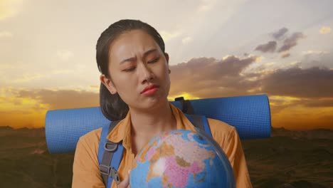 primer plano de una excursionista asiática con una mochila de montañismo mirando el globo mundial en sus manos y haciendo un gesto confuso mientras está de pie en la cima de la montaña durante la hora de la puesta del sol