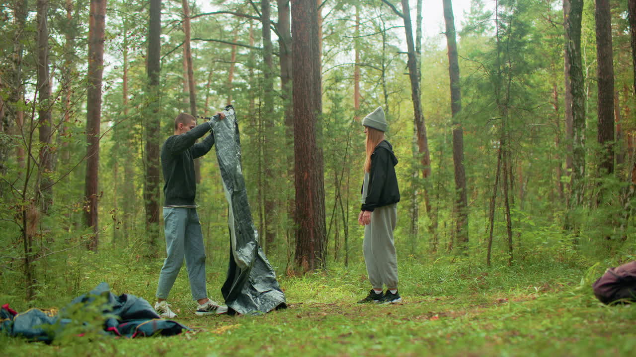 Young boy lifts and opens tent fabric while lady prepares ground for setup in forest, working together to spread material on grass among tall trees and scattered bags