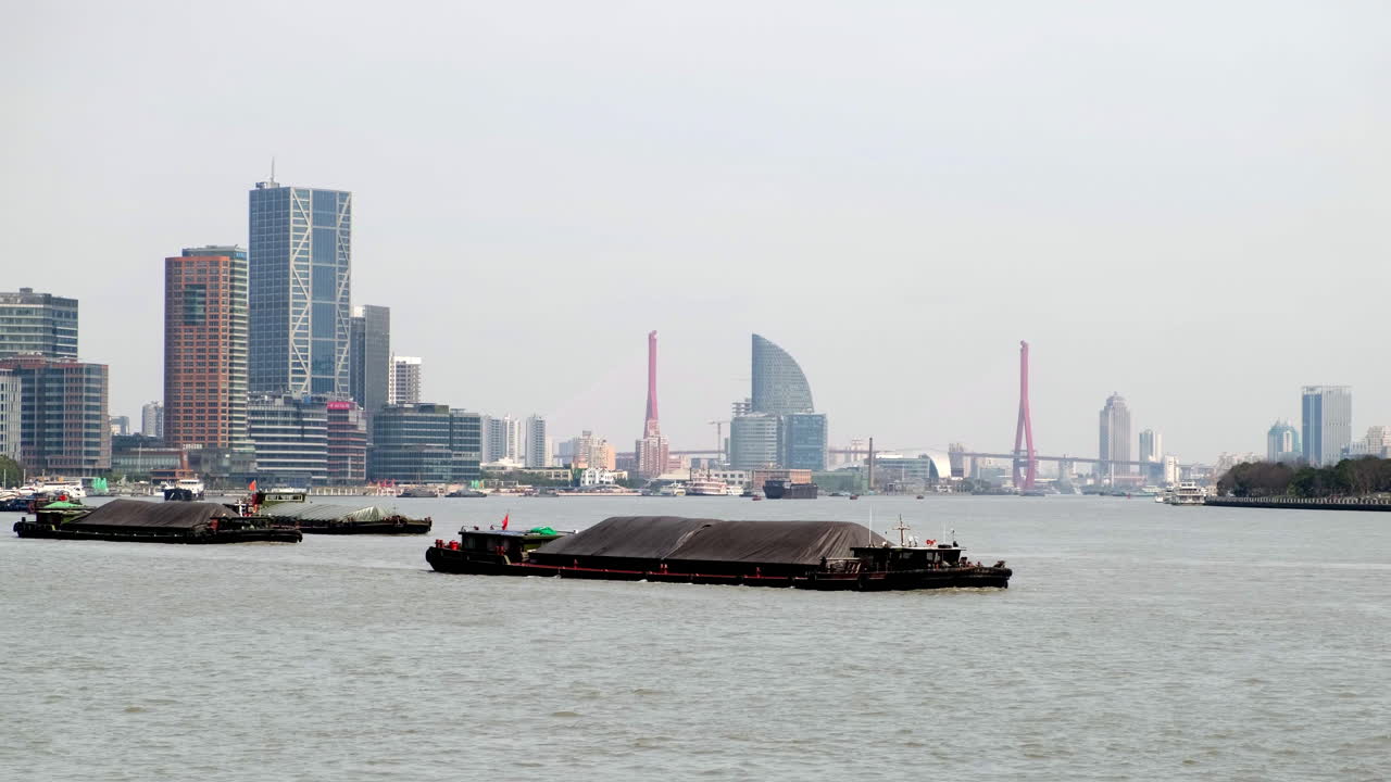 Long barges on Huangpu River in Shanghai, with city skyline and iconic bridges in the hazy distance