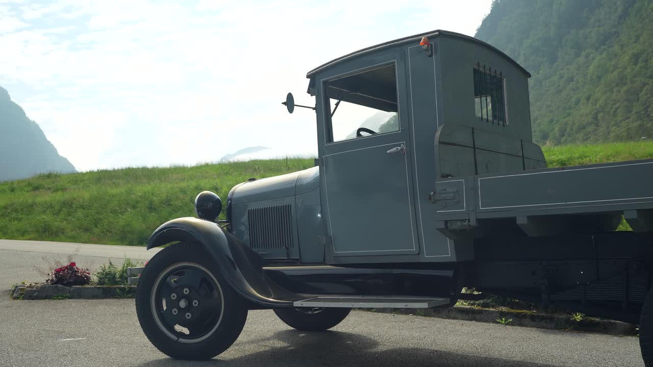 A classic gray truck is parked along a rural road surrounded by lush green fields and towering mountains under a partly cloudy sky. The vehicle showcases vintage design elements