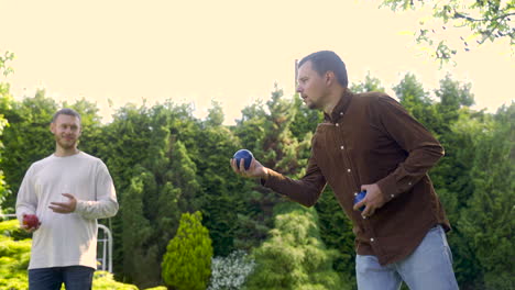 side view of caucasian young man throwing a blue petanque ball in the park on a sunny day while his male friend waits his turn