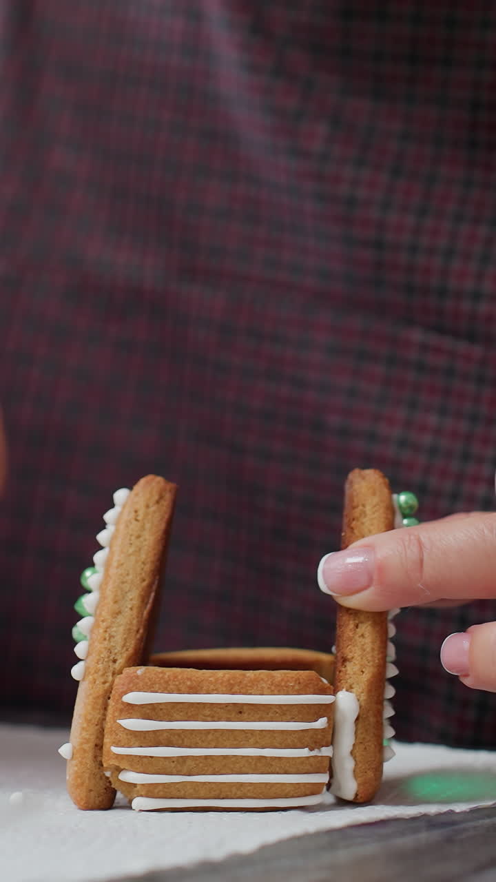 집 모양의 디자인을 형성하기 위해 진저브레드 쿠키를 장식하기 위해 아이싱을 사용하는 베이커의 클로즈업, 부 배경에 꽃 쿠키와 다른 베이킹 필수품의 흐릿한 시각