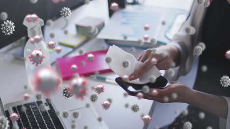 animación de células de covid-19 flotando sobre una mujer limpiando un teléfono inteligente en la oficina